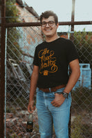 Male model standing and wearing out Reel To Reel t-shirt. Black short sleeve t-shirt with orange cursive letters that says The Next Waltz. The words are coming out of an orange vintage tape player at the bottom of the shirt.