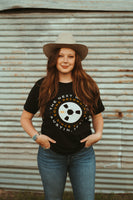 Female model standing and wearing out Reel Wheel T-shirt. Black short sleeve t-shirt with white and orange letters that go around in a circle that says The Next Waltz Sound Recordings Since 2017 Austin, TX. In the center of the shirt there is a cream colored tape wheel with The Next Waltz logo in orange. The words circle around the tape reel.
