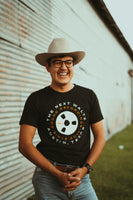 Male model standing and smiling wearing our Reel Wheel Tee.Black short sleeve t-shirt with white and orange letters that go around in a circle that says The Next Waltz Sound Recordings Since 2017 Austin, TX. In the center of the shirt there is a cream colored tape wheel with The Next Waltz logo in orange. The words circle around the tape reel.