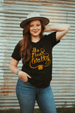 Female modeling wearing a cowboy hat and standing up and wearing our Reel To Reel t-shirt. Black short sleeve t-shirt with orange cursive letters that says The Next Waltz. The words are coming out of an orange vintage tape player at the bottom of the shirt.