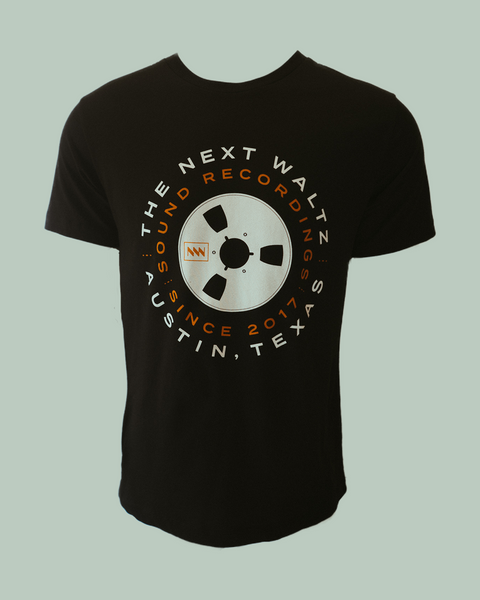 Black short sleeve t-shirt with white and orange letters that go around in a circle that says The Next Waltz Sound Recordings Since 2017 Austin, TX. In the center of the shirt there is a cream colored tape wheel with The Next Waltz logo in orange. The words circle around the tape reel.